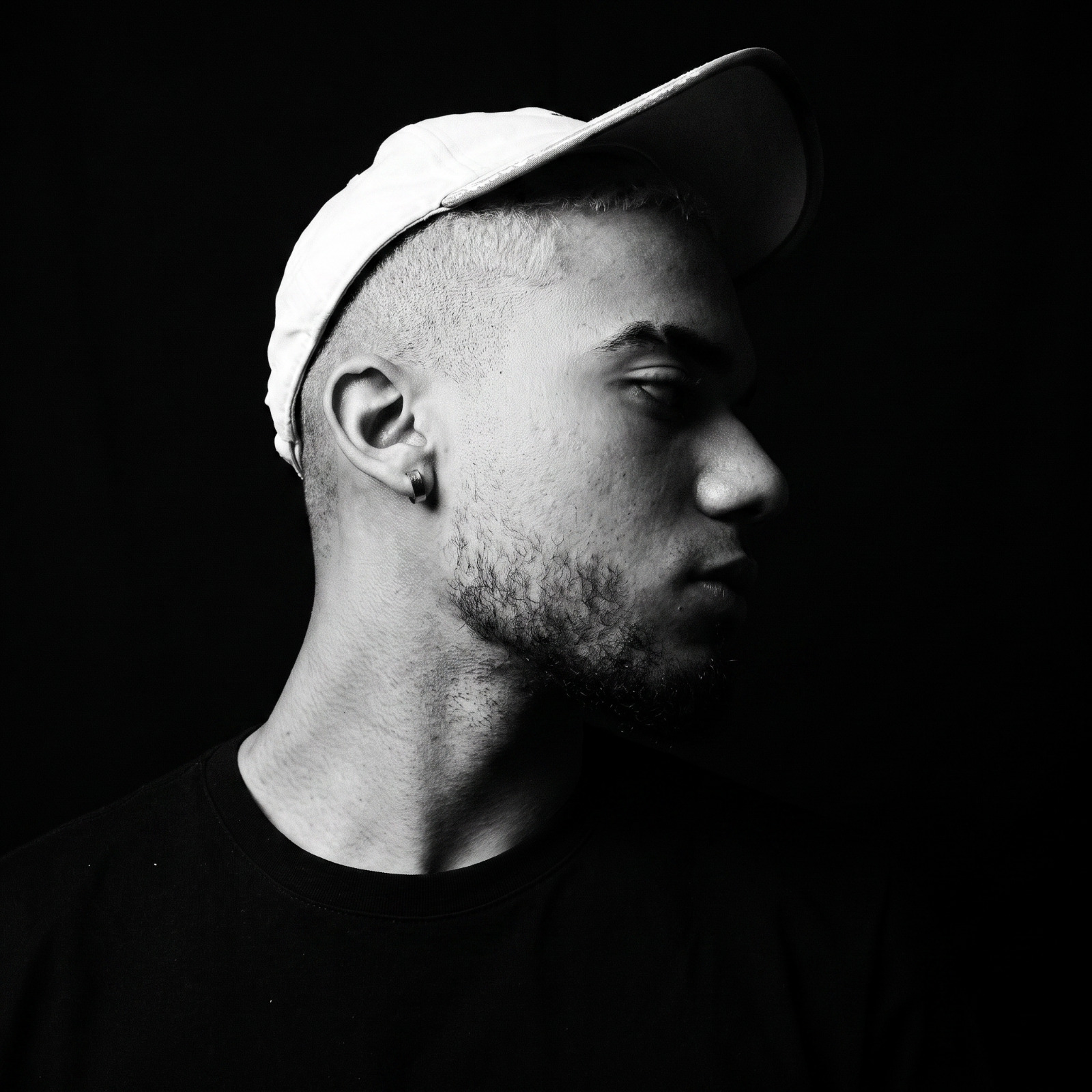 Retrato en blanco y negro de Raleey de perfil, con gorra clara, sobre fondo negro